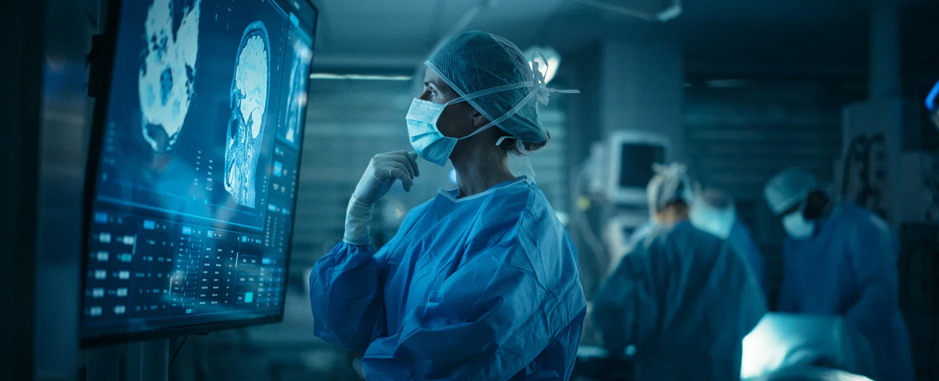 A medical professional in scrubs and gloves examines brain scans displayed on a large screen in a surgical environment.