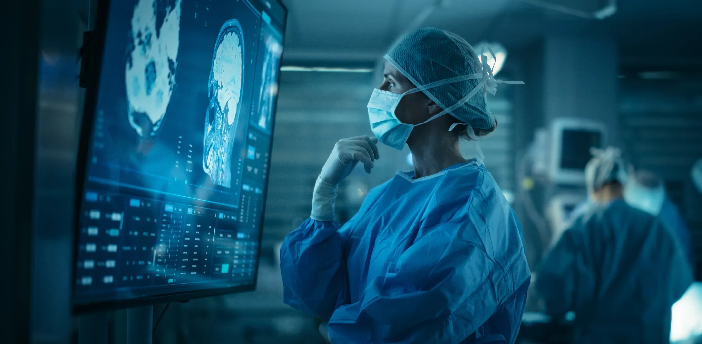 A medical professional in scrubs and gloves examines brain scans displayed on a large screen in a surgical environment.
