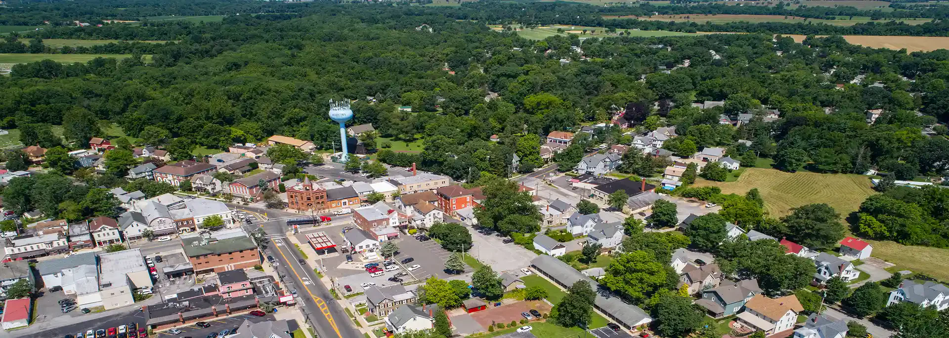 Aerial view of a small town surrounded by lush greenery, featuring a water tower and residential buildings interspersed with commercial properties.